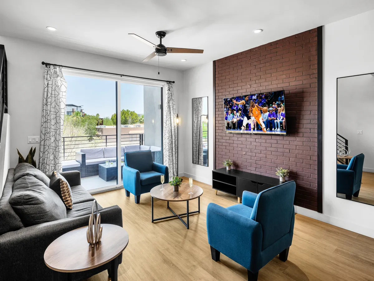 The image showcases a spacious, sunlit living room with plush blue chairs, a comfortable sofa, a stylish coffee table, and a large TV displaying a live sports game. The setup invites guests to gather, relax, and enjoy lively moments together.