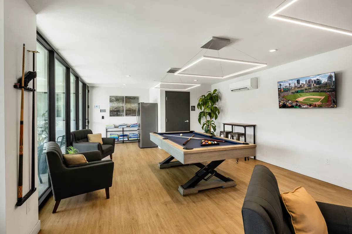 A vibrant interior clubhouse scene featuring a pool table at the center, surrounded by plush sofas, a wall-mounted TV, and lush greenery. The space exudes an atmosphere perfect for friendly games, sports viewing, and relaxed social gatherings.