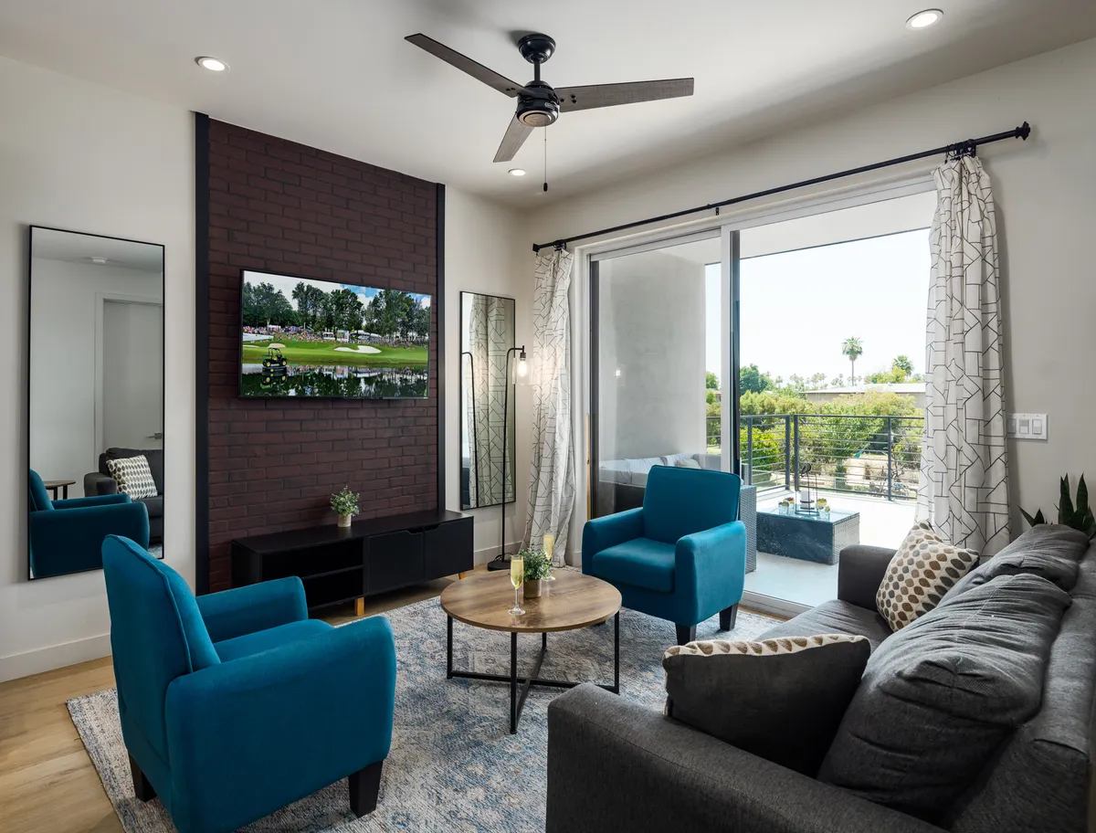 A comfortable living area with a sofa, chairs, coffee table, smart TV, and sliding glass doors opening to a private balcony. The room is filled with natural light and invites both relaxation and seamless indoor-outdoor living.