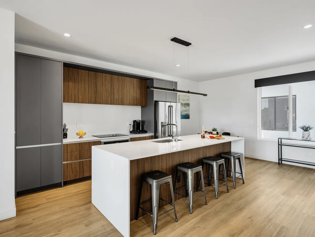 A contemporary kitchen with an island countertop, stainless steel appliances, wood cabinetry, and bar stools. The space is open and designed for both meal preparation and casual socializing.