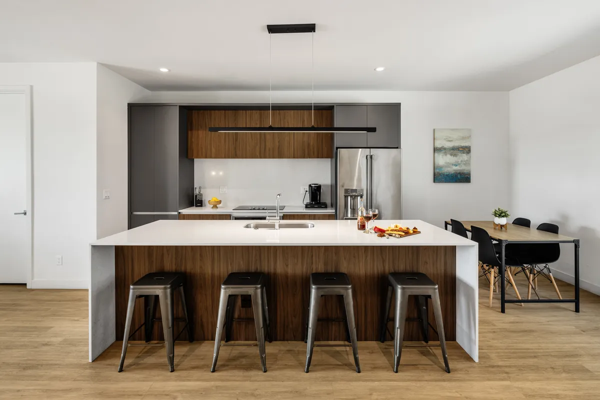 A modern kitchen space showcasing a large kitchen island with bar stools, a dining table nearby, art on the wall, and high-end appliances. The design invites guests to cook, dine, and socialize in an open, stylish setting.