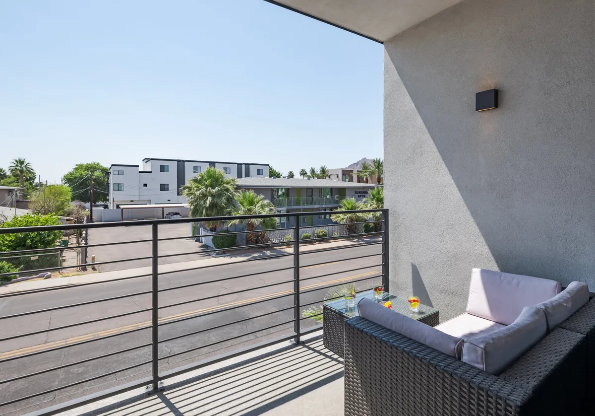 This image shows a balcony seating area overlooking a street lined with palm trees. The view captures the outside atmosphere, emphasizing the connection to the neighborhood and community. Comfortable outdoor chairs invite guests to sit and relax, highlighting the opportunity for socializing or enjoying quiet moments outside the private condos.