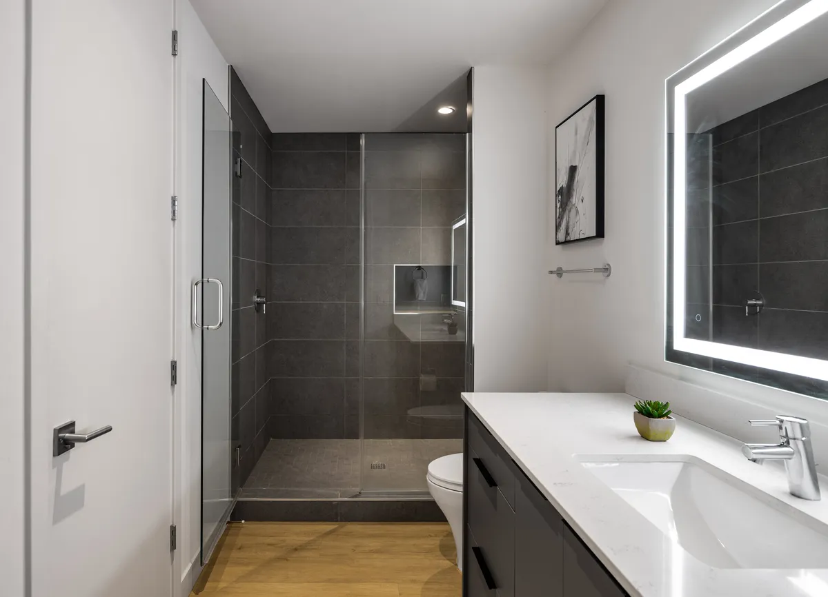 A contemporary bathroom scene with a glass shower enclosure, a modern sink and vanity, an illuminated mirror, and decorative wall art. The space is bathed in soft, even lighting, conveying a spa-like ambiance ideal for personal grooming and self-care routines.