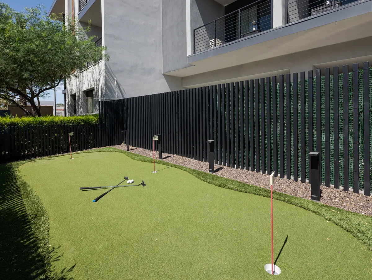 A playful exterior view of a putting green surrounded by modern architectural elements and fencing, with golf clubs ready for friendly competitions. The outdoor space is sunny and designed for leisurely fun and social gatherings.