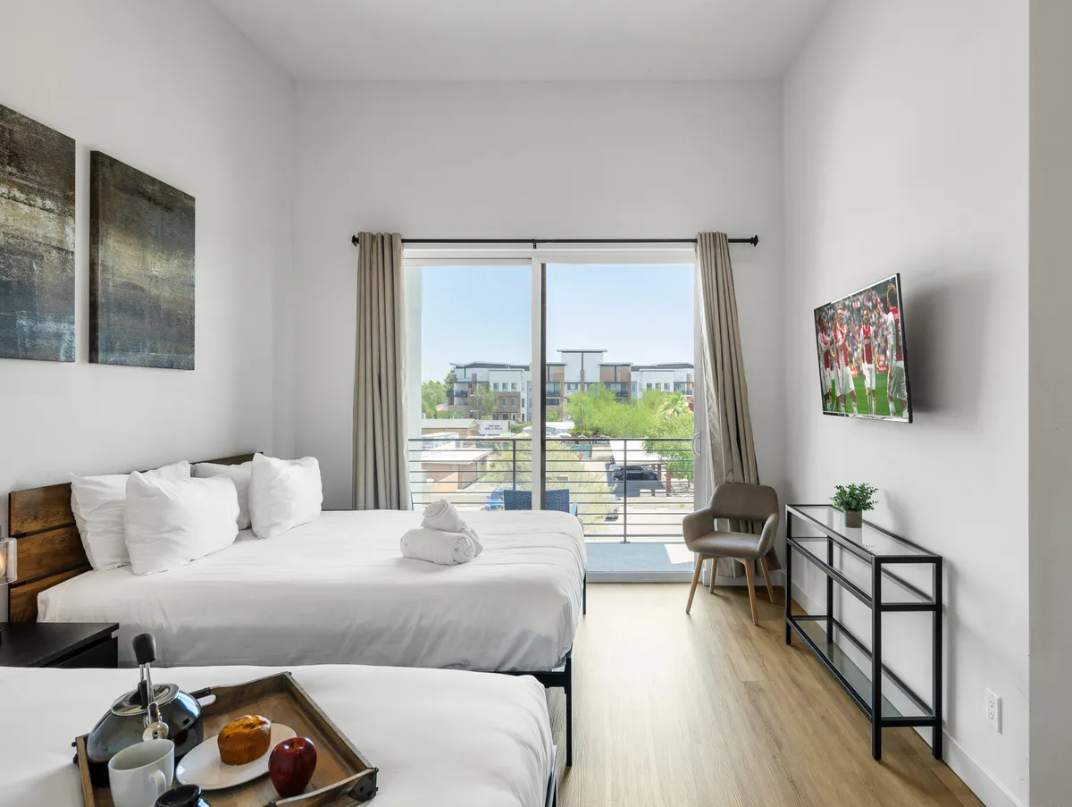 A bright, modern bedroom with two beds dressed in crisp white linens, a large window leading to a balcony, a TV mounted on the wall, and decorative artwork. The room is filled with natural light, creating a welcoming space for rest and privacy.