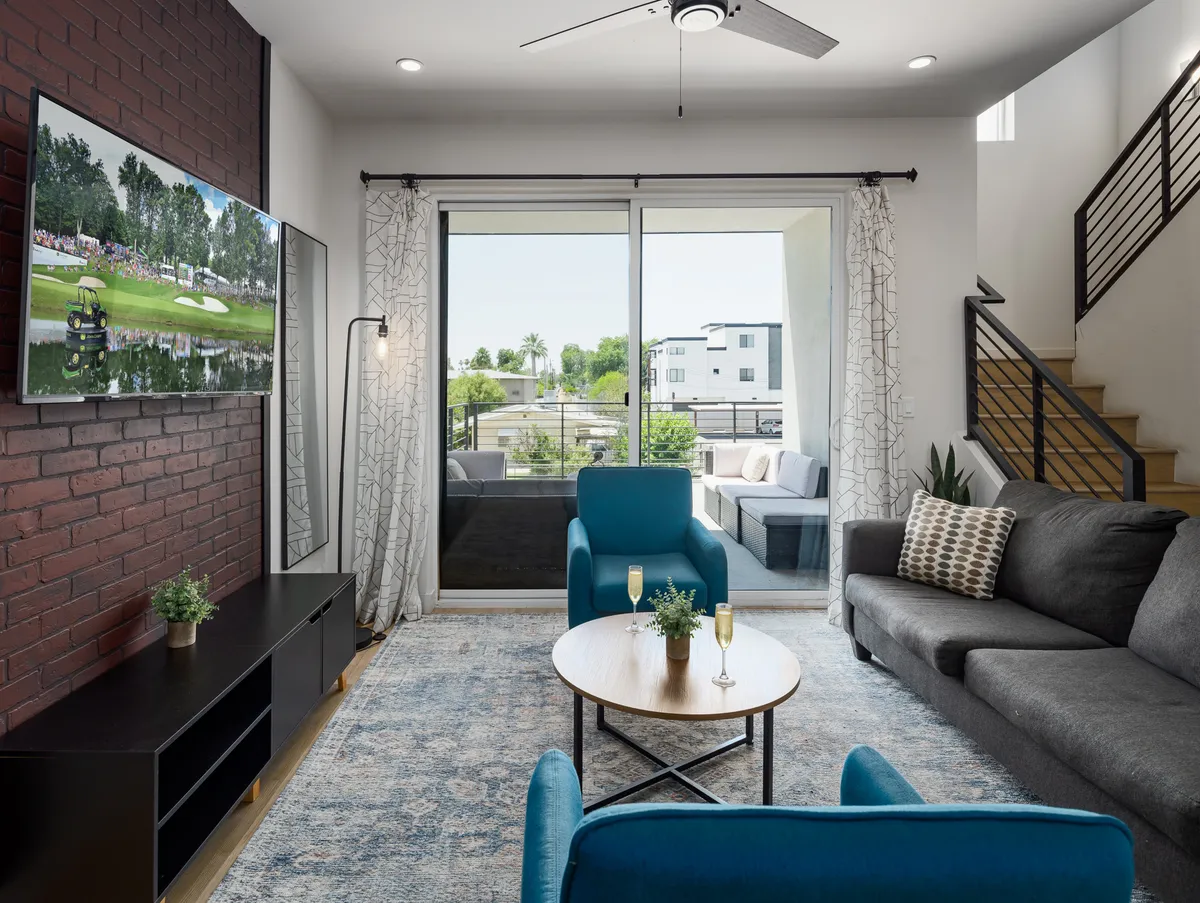 A stylish living room featuring bright blue chairs, a coffee table with drinks and plants, a large TV showing a sports event, and a sliding glass door that opens to a balcony. The atmosphere is lively and social.