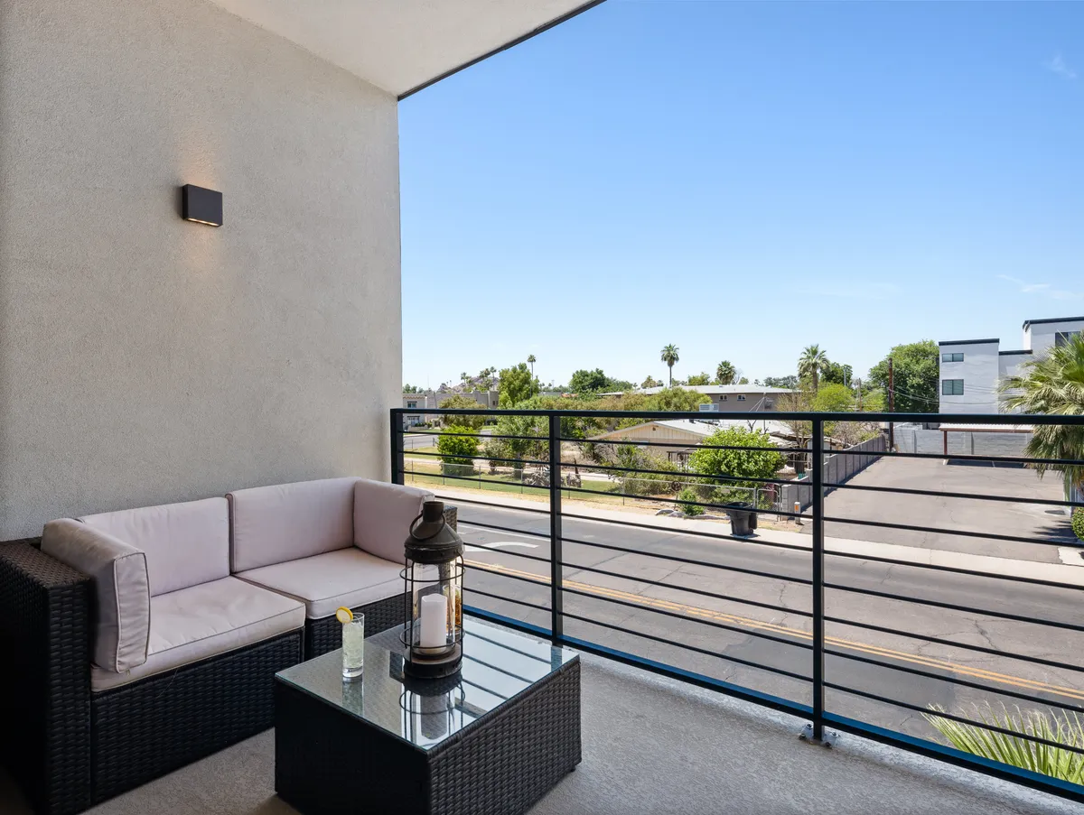 A tranquil exterior shot showing a private balcony setup with comfortable seating, a glass table, and a lantern, ideal for relaxing or socializing. The setting evokes the feeling of unwinding outdoors, with gentle sunset lighting creating an inviting atmosphere.
