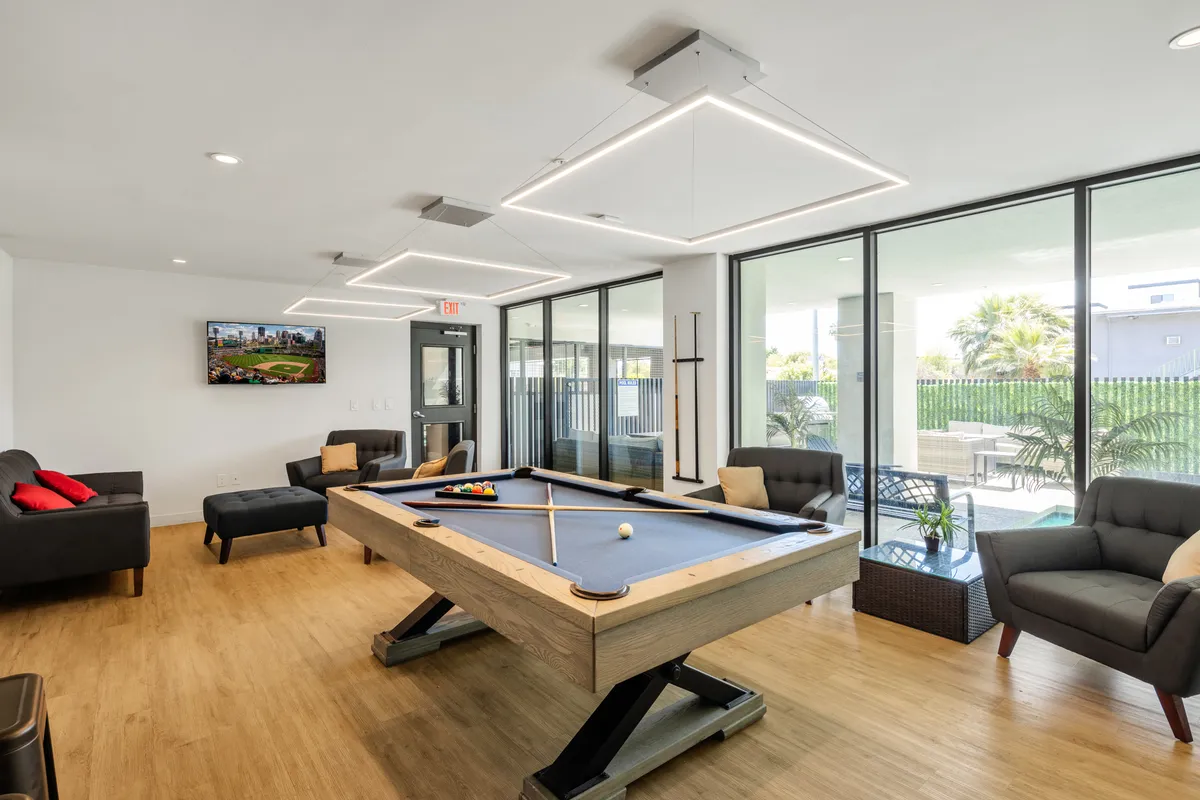 A spacious clubhouse interior featuring a pool table, cozy sofas, a television, and large windows that let in natural light. The room is designed for friendly competition, group entertainment, and relaxed socializing.