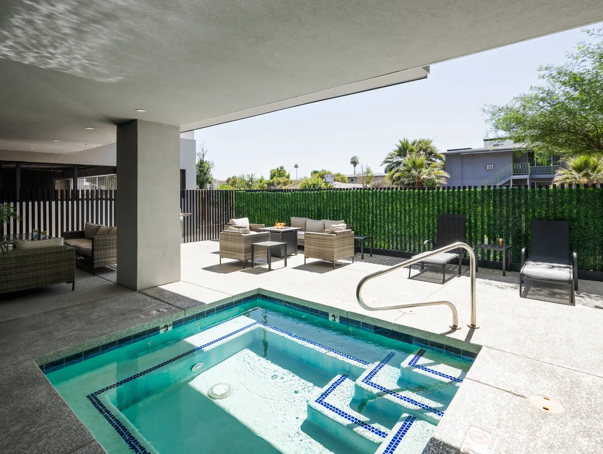 An inviting outdoor setting with a bubbling hot tub, comfortable seating around the area, lush landscaping, and a green privacy wall. The tranquil scene is set for relaxation and socializing, particularly during sunset.