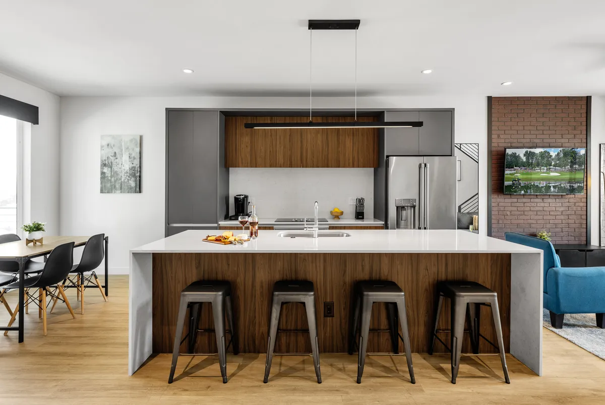 A contemporary open-concept living and kitchen area featuring a spacious kitchen island with stools, modern appliances, a dining area, and a smart TV visible from both the kitchen and living space. The open layout encourages easy flow between cooking, dining, and relaxing.